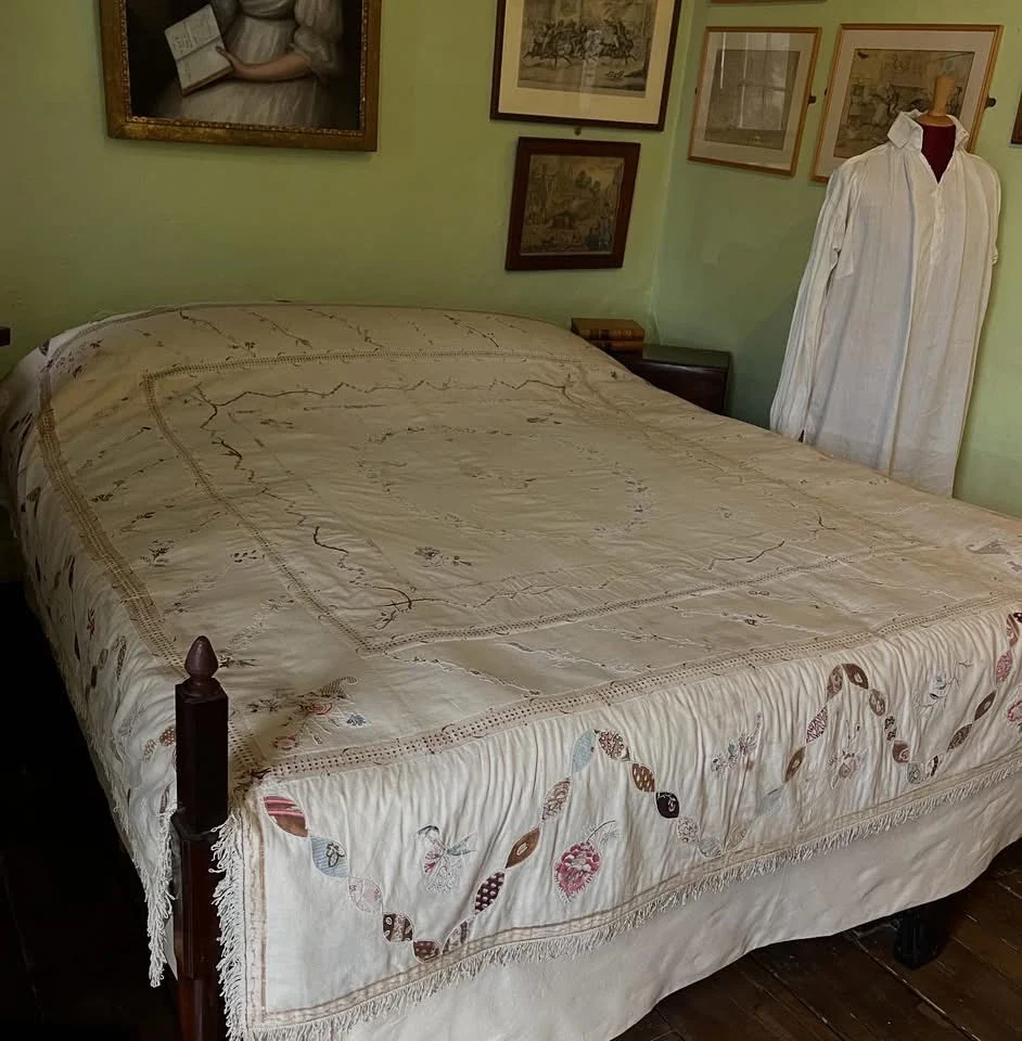 A bed covered in a historic patchwork counterpane in a museum setting with framed prints and paintings on the green painted walls, alongside a mannequin wearing a plain white nightshirt
